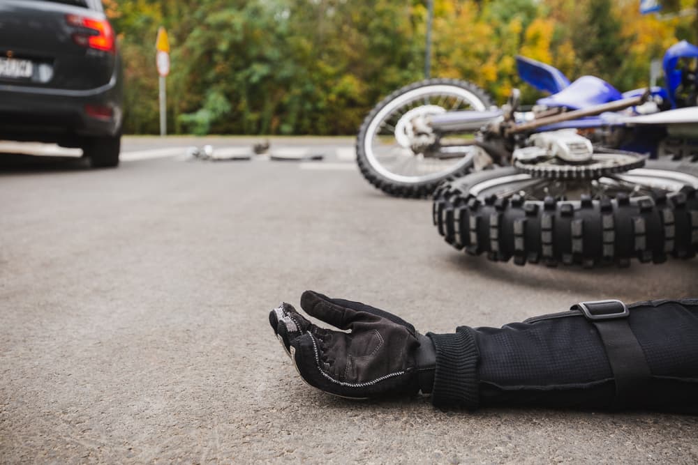 A gloved arm on the roadway beside a fallen motorcycle and a car.