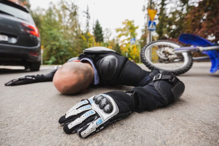 A motorcyclist lies on the road beside a fallen blue motorcycle and a car.