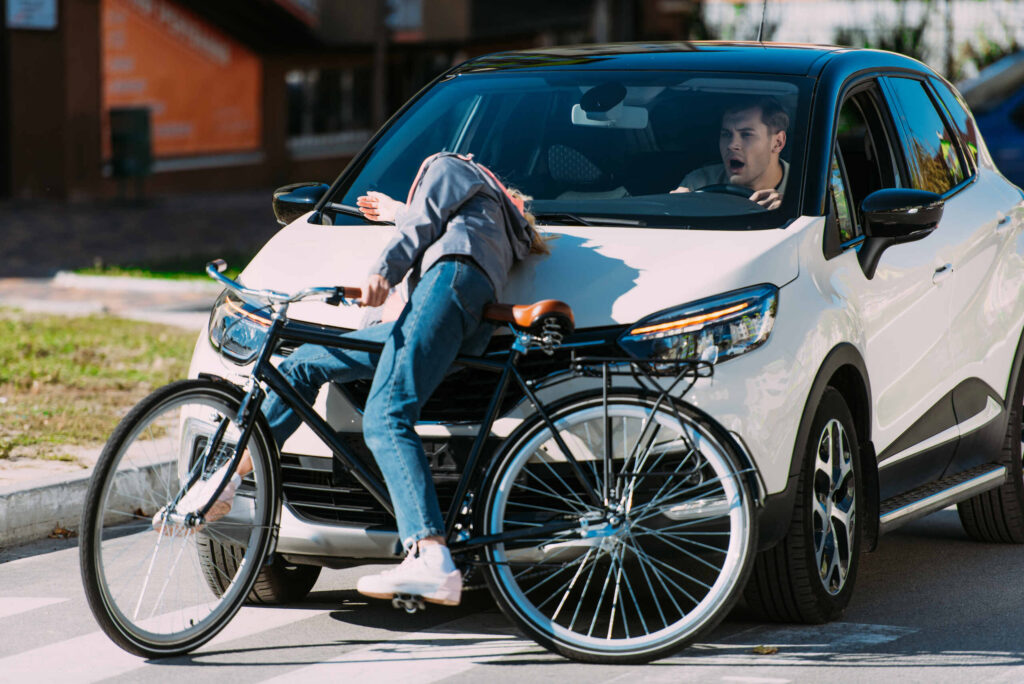 A white car colliding with a cyclist on a city street, showing the rider falling onto the hood while the driver looks on in shock.