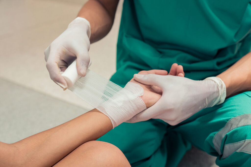 A medical professional in green scrubs and white gloves carefully applies a gauze bandage to a patient's burned hand and wrist