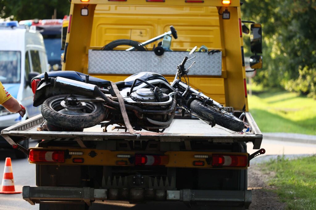A damaged, wrecked motorcycle strapped down flat onto the flatbed of a yellow tow truck at a roadside incident scene, illustrating the aftermath of crashes handled by Broward County motorcycle accident lawyers.