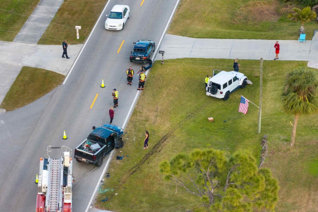 An aerial view of a multi-vehicle car accident scene on a residential road, showing emergency responders, a fire truck, a damaged pickup truck, and a white Jeep on the grass, illustrating incidents handled by Broward County personal injury lawyers.