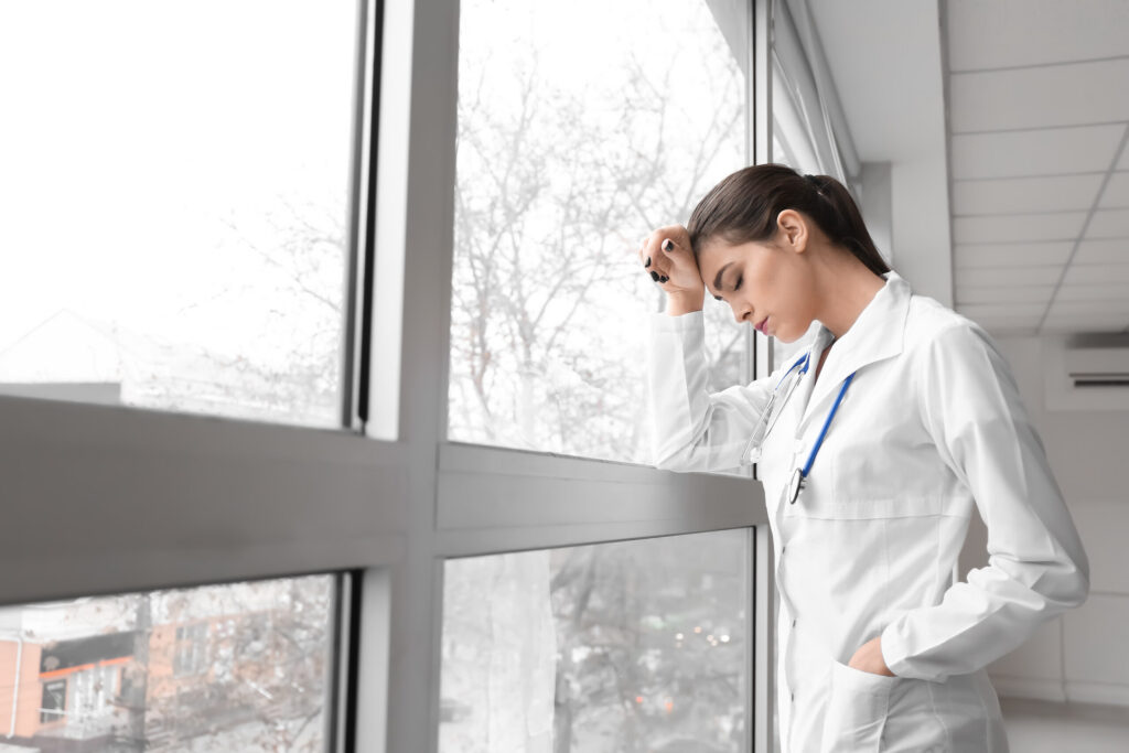 A stressed female doctor in a white lab coat leans against a large hospital window with her head in her hand, representing medical negligence cases evaluated by Broward County wrongful death lawyers.