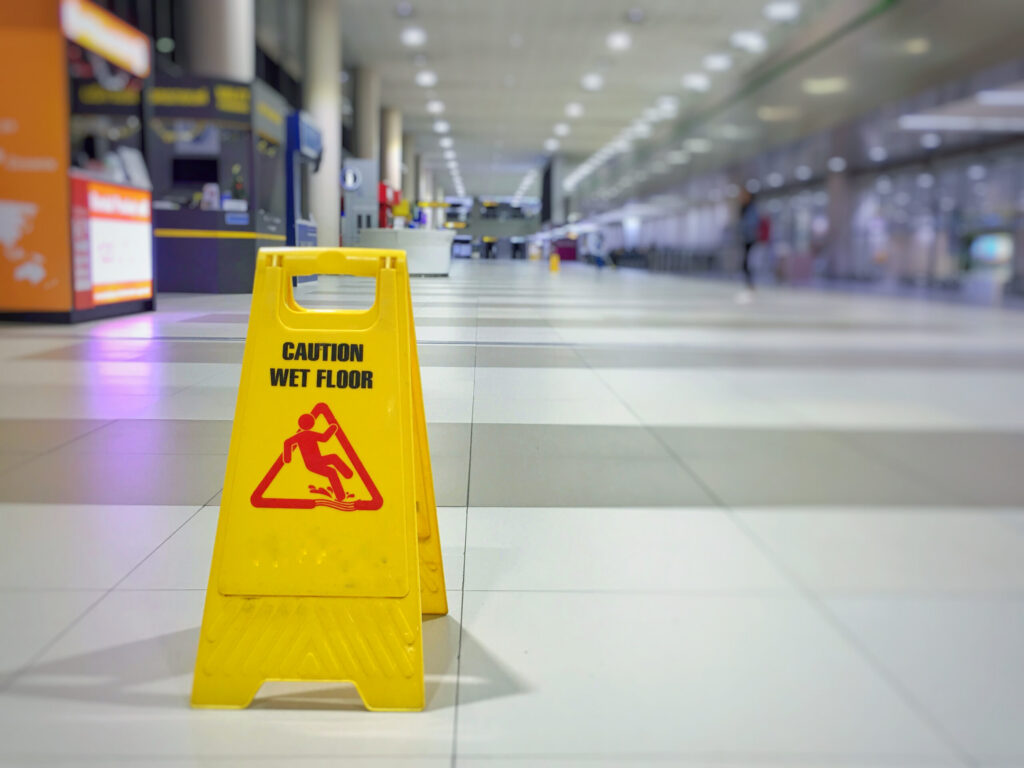 A bright yellow "Caution Wet Floor" sign stands on a polished tile walkway inside a large, blurred public terminal or building, highlighting hazards evaluated by Broward County slip and fall lawyers.
