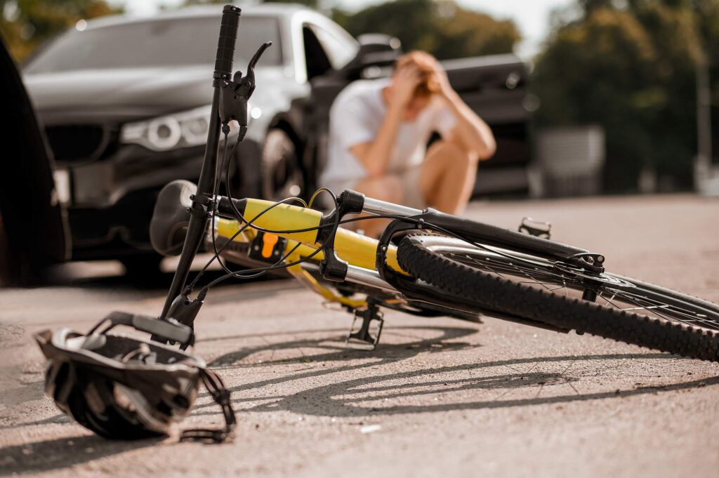 A yellow bicycle and helmet lie on the asphalt in front of a black car with a distressed person sitting in the background, representing cases handled by Broward County Bicycle Accident Lawyers.