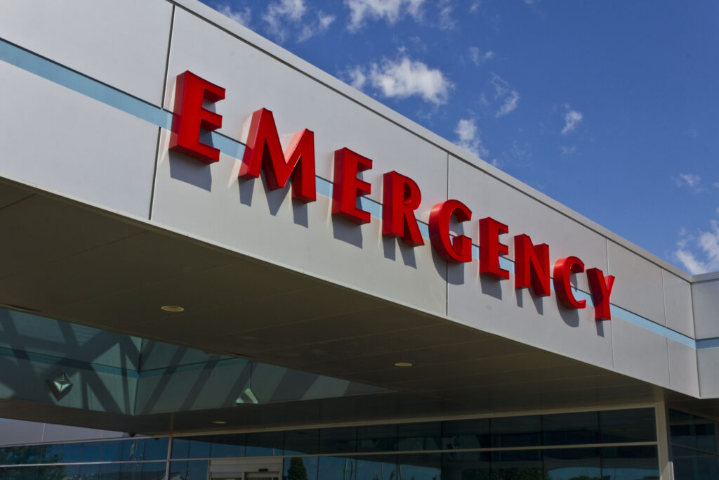 Large red "EMERGENCY" sign on the exterior facade of a hospital building under a blue sky, representing urgent care for cases handled by Broward County Catastrophic Injury Lawyers.