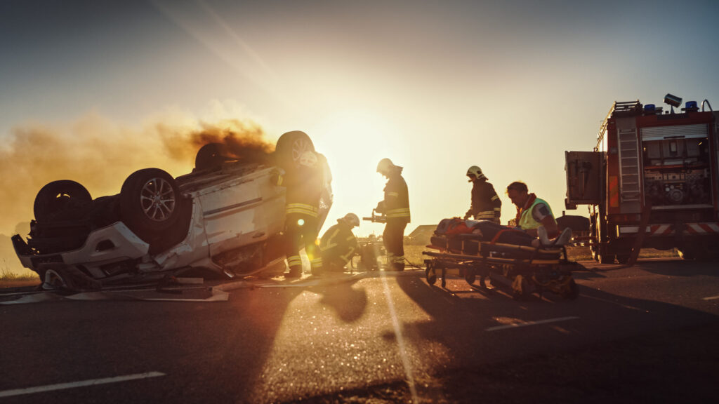 First responders and firefighters attending to a severe car crash with an overturned vehicle at sunset, illustrating the type of severe accidents managed by Broward County Catastrophic Injury Lawyers.