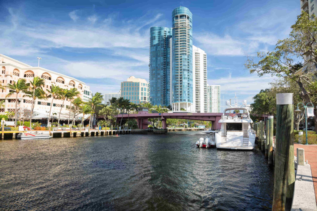 A scenic view of Fort Lauderdale's Riverwalk area featuring a canal with boats, palm trees, and tall modern glass high-rises under a blue sky, establishing the local setting for claims involving a drunk driver in Broward County.