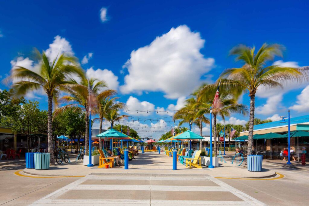 A sunlit, outdoor pedestrian plaza in Broward County lined with palm trees, patio umbrellas, and colorful chairs, providing geographic context for Broward County Spinal Cord Injury Lawyers.
