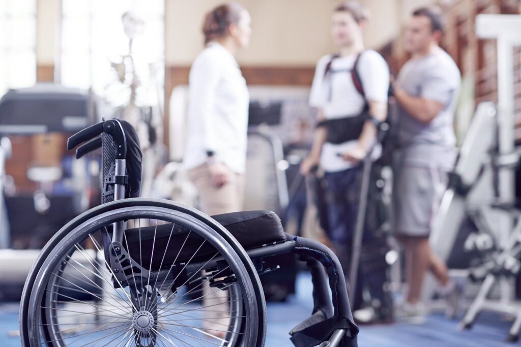 A close-up of an empty wheelchair in a rehabilitation clinic, with a patient and medical team blurred in the background, representing cases handled by Broward County Spinal Cord Injury Lawyers.