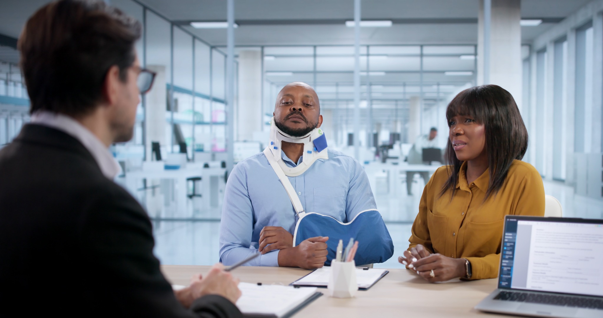 A man wearing a neck brace and arm sling sits beside a woman during a legal consultation