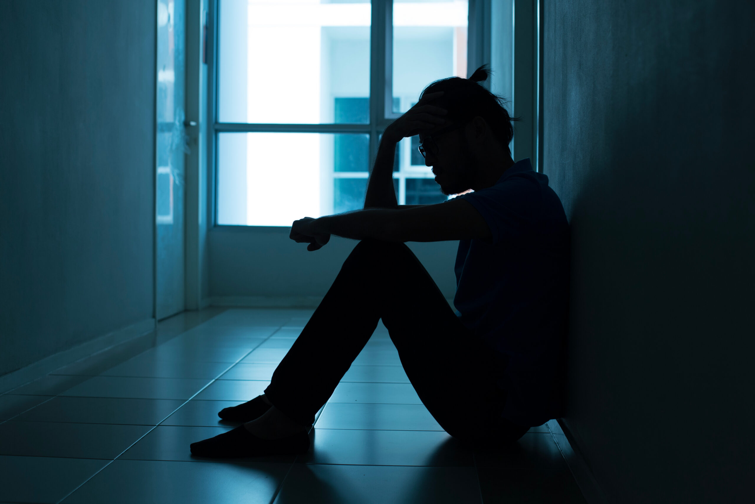Silhouette of a man sitting on the floor beside a window, resting his head in one hand.